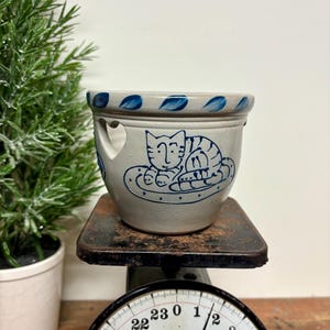 May include: A light gray stoneware crock featuring a blue hand-painted cat illustration on the front, a blue leaf-patterned rim, and heart-shaped cutouts on the sides. The crock is positioned on a vintage scale with a dial marked 24 Lbs.