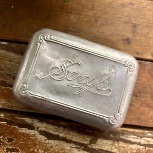 May include: A rectangular, silver-colored soap dish with the word "Soap" in cursive script on the lid. The dish has an ornate border design and a slightly weathered appearance, suggesting it is vintage.