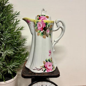May include: White porcelain teapot with a floral rose design in pink and green. The teapot has a yellow rim and a decorative handle. It is displayed on a vintage scale.