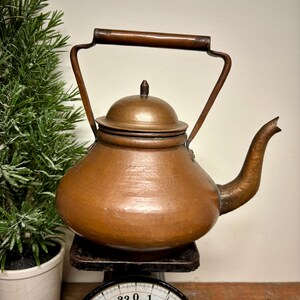 May include: A vintage copper kettle with a dark wooden handle and spout. The kettle sits on a black scale with a white face and black numbers. A small green plant in a white pot is to the left of the kettle.