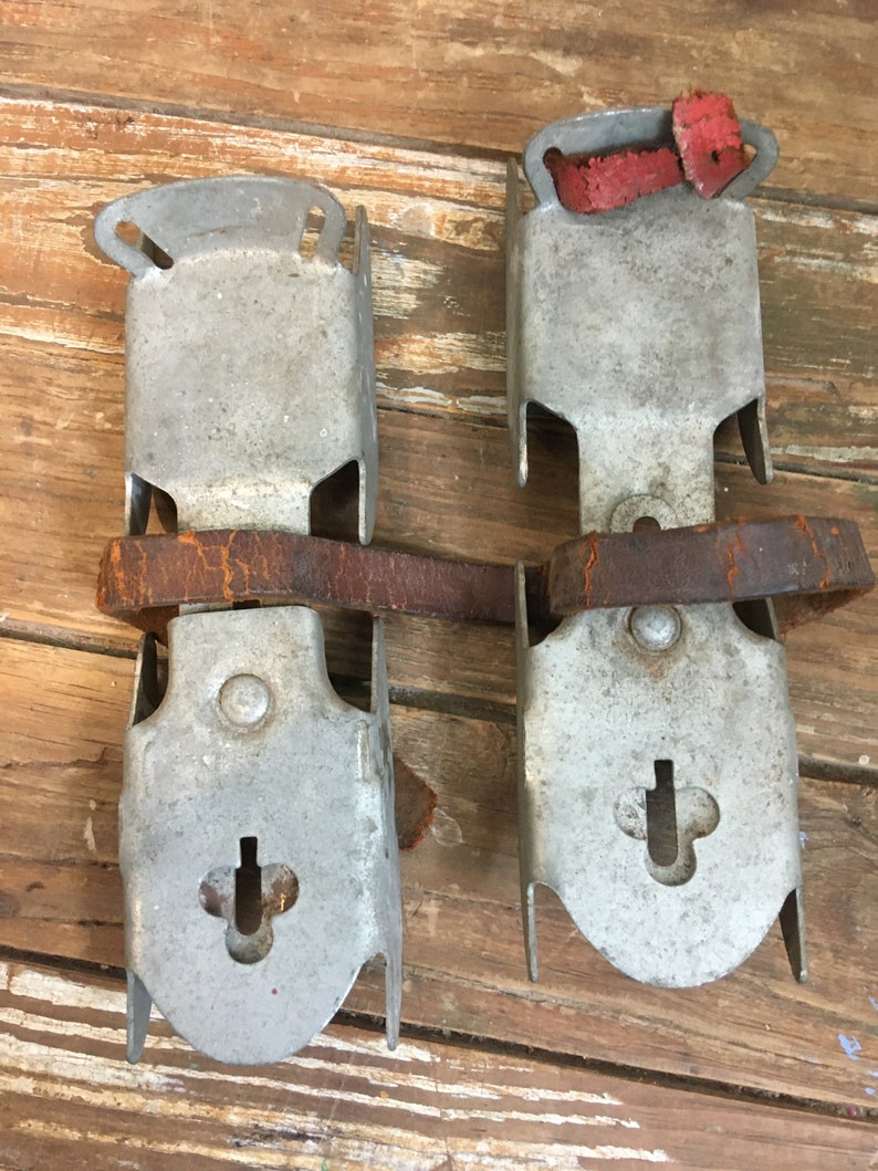 May include: A pair of vintage metal ice skate attachments with leather straps and red rubber pads. The metal is silver and shows signs of wear.
