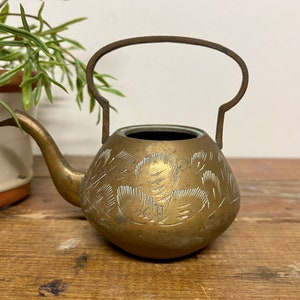 May include: A small, brass teapot with an ornate design. The teapot has a handle and a spout, and is decorated with a floral pattern.