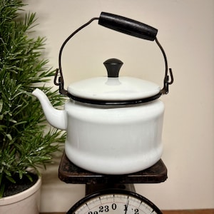 May include: A white enamelware tea kettle with a black handle and lid knob. The kettle sits on a vintage scale. The scale reads 24 lbs. 0 oz. and has a black and white face. A small green plant in a white pot is to the left.