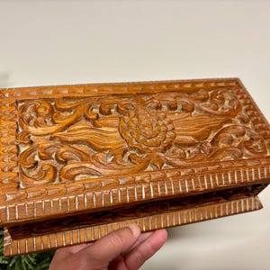 May include: A rectangular wooden box with intricate floral carvings. The box is a warm brown color and features detailed relief patterns on the lid and sides. The box is held by a hand.