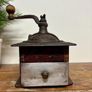 May include: Antique coffee grinder with a wooden handle and a metal crank. The grinder has a red and black painted wooden body with a metal drawer. The metal components show signs of age and wear, adding to its vintage appeal.