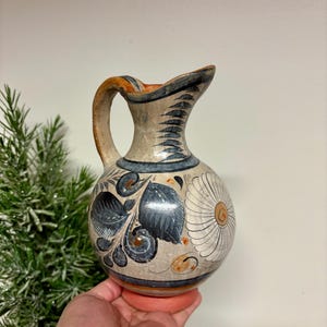 May include: Hand-painted ceramic pitcher with a rounded body and a handle. The pitcher features a cream-colored base with blue and brown floral designs, a white flower, and a blue rim. The pitcher is held by a hand.