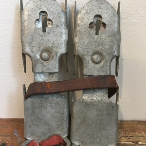 May include: A pair of vintage metal ice skate attachments with a brown leather strap and red fabric straps. The attachments have a clover-shaped hole in the top.