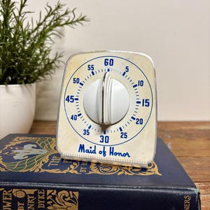 May include: A vintage Maid of Honor kitchen timer with a white face, blue numerical markings from 5 to 60, and a central white dial knob. The timer is rectangular with rounded corners and features a metallic silver finish on the outer edges.