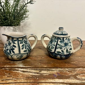 May include: A cream-colored creamer and covered sugar bowl set with blue floral designs. The ceramic pieces have a crackle glaze and curved handles. A small plant in a white pot is in the background.