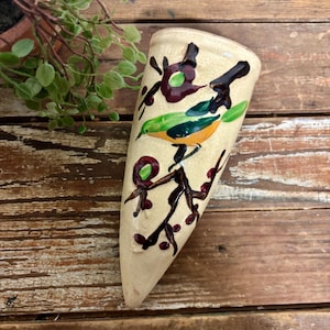 May include: A cream-colored, ceramic wall pocket vase with a painted bird and branch design. The bird is green, orange, and blue, perched on a brown branch with maroon accents. The vase is approximately 8 inches tall.