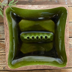 May include: A green ceramic ashtray with a wood-grain rim. The ashtray has five indentations for cigarettes.