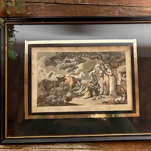 May include: Framed antique print with a black and gold frame. The print depicts a pastoral scene with figures, animals, and a building. The artwork is set against a cream-colored mat.