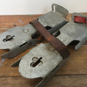 May include: A pair of vintage metal ice skate attachments with leather straps. The straps are brown and have red leather accents. The metal is silver and has a worn look.