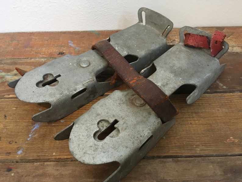 May include: A pair of vintage metal ice skate attachments with leather straps. The straps are brown and have red leather accents. The metal is silver and has a worn look.