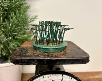 May include: A circular green metal flower frog featuring a series of upright, thin wire prongs arranged in a dense, radiating pattern. The base is painted green with visible signs of wear and oxidation, showing a weathered, aged texture.
