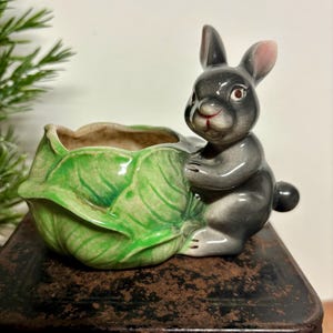 May include: A ceramic planter featuring a gray bunny holding a green cabbage-shaped container. The bunny has pink-lined ears and red eyes. The planter is a decorative item, likely for small plants or succulents.