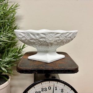 May include: A white, footed dish with a floral design. The dish is made of ceramic and sits on a vintage scale. The dish has a scalloped edge and is decorated with embossed flowers and leaves. The scale is dark brown and has a black face.