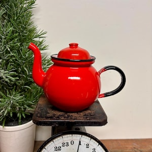 May include: A vibrant red enamel teapot with a black handle and rim sits on a vintage scale. The teapot has a rounded body and a curved spout. The scale reads 24 lbs. 0 oz. A small green plant is in the background.