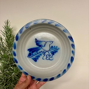 May include: A round ceramic bowl with a light gray speckled finish. The center features a blue hand-painted design of a bird perched on a leafy branch with a small flower. The rim is decorated with a repeating pattern of blue painted leaf shapes.