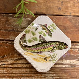May include: A white, diamond-shaped decorative plate featuring two colorful fish illustrations. The fish are green, yellow, and pink, with detailed scales and fins. The plate has a textured, white border and is set against a rustic wooden surface.