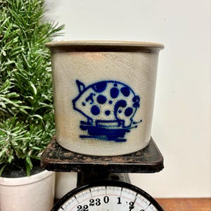 May include: A stoneware crock with a blue pig design. The cylindrical container has a tan and brown color scheme. The pig illustration features a series of blue spots. The crock is sitting on a vintage scale.