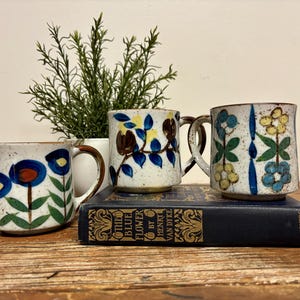 May include: Three ceramic mugs with a speckled off-white glaze and hand-painted floral designs in blue, yellow, and green. The mugs are arranged on a dark blue book titled The Blue Flower by Henry Van Dyke, with a green plant behind them.