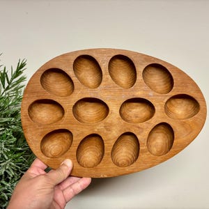 May include: A wooden egg tray with 12 oval-shaped indentations. The tray is made of light brown wood and is held by a person's hand. The tray is designed to hold eggs and is a kitchen accessory.