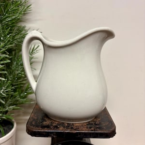 May include: A solid off-white ceramic pitcher with a curved handle and a pouring spout. The pitcher is positioned on a vintage metal scale with a circular dial showing numbers, including 21, 22, 23, 0, 1, 2, and 3, with a black metal platform.