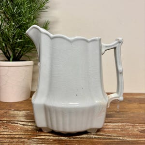 May include: A light blue ceramic pitcher featuring a scalloped rim, a vertical ribbed base, and a textured handle. The surface of the pitcher displays a distinct crackle glaze finish throughout, with subtle variations in the blue tone.