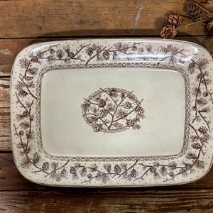 May include: A rectangular ceramic serving platter with a cream-colored base and a brown transferware pattern. The design features a central oval motif of pine branches and cones, surrounded by a border of pine needles and pinecones.