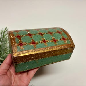 May include: A small, antique wooden chest with a curved lid. The chest is painted in shades of green and gold, with a decorative pattern of red and gold shapes on the lid. The chest is held in a person's hand.
