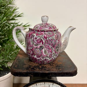 May include: A vintage teapot with a floral pattern in shades of pink, white, and gray. The teapot has a white handle and spout with gold trim. The teapot is sitting on a rustic, dark metal surface.