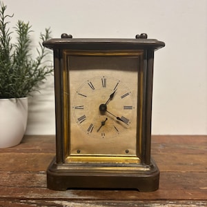May include: A rectangular mantel clock with a dark bronze-toned metal frame and a gold-toned inner border. The clock face is off-white with black Roman numerals, black hands, and a smaller sub-dial at the bottom for seconds. It has a flat top.