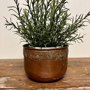 May include: A round copper planter with a weathered patina and a decorative embossed band around the upper rim. The planter contains a lush, artificial green plant with thin, needle-like leaves that extend upwards from the center.