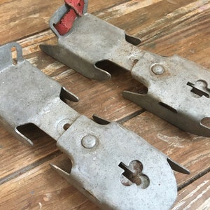 May include: A pair of vintage metal ice skate attachments with red leather straps. The attachments have a clover-shaped hole in the center.