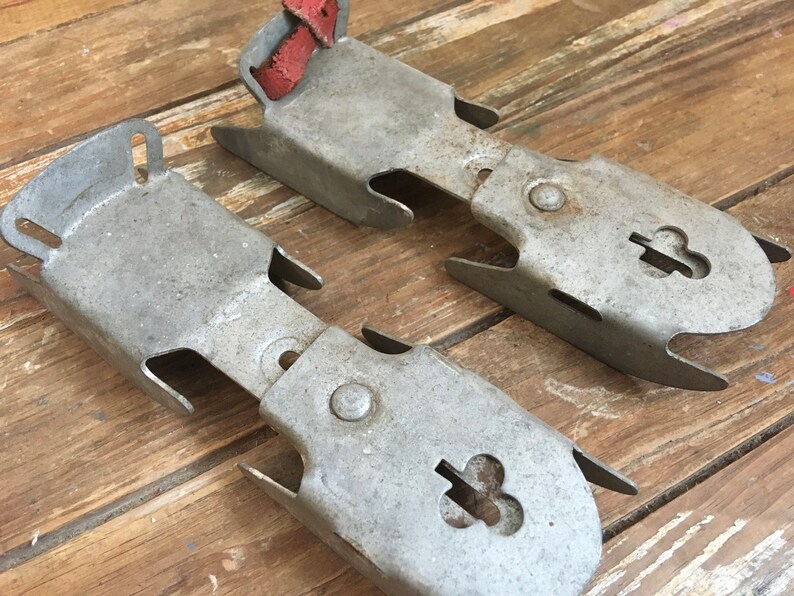 May include: A pair of vintage metal ice skate attachments with red leather straps. The attachments have a clover-shaped hole in the center.