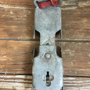 May include: A vintage metal ice skate holder with a red leather strap. The holder has a keyhole and a small round hole. The holder is in good condition with some signs of wear.