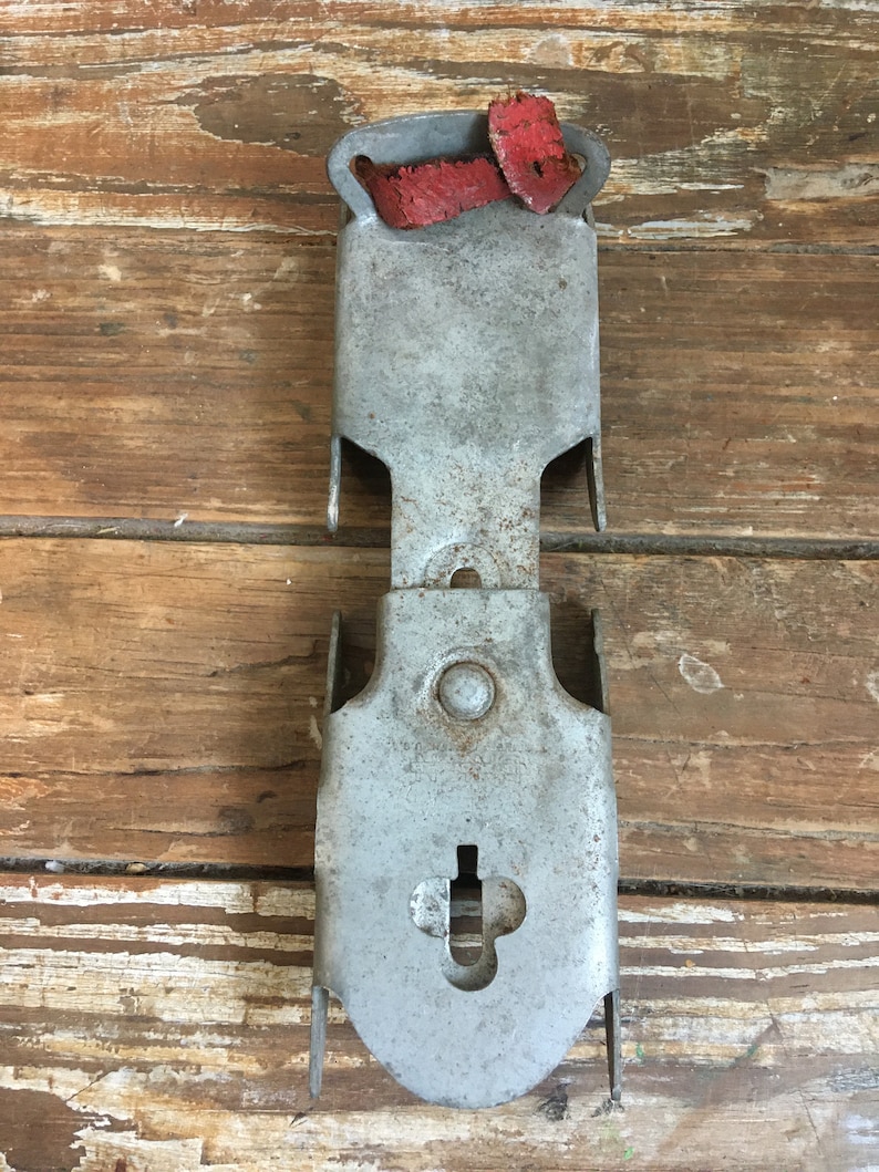 May include: A vintage metal ice skate holder with a red leather strap. The holder has a keyhole and a small round hole. The holder is in good condition with some signs of wear.