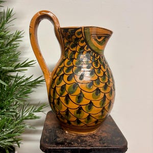 May include: A ceramic pitcher with a handle, painted in shades of orange, green, and black. The pitcher features a fish scale pattern and a curved handle. The pitcher is sitting on a black surface.