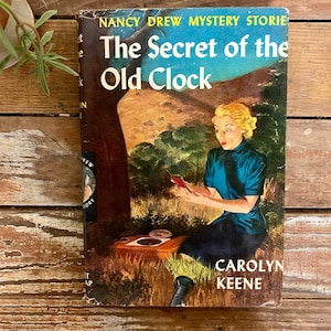 May include: A vintage paperback book cover featuring a young woman in a blue dress holding a small red object. The book is titled "The Secret of the Old Clock" and is part of the Nancy Drew Mystery Stories series by Carolyn Keene.