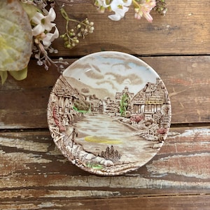 May include: A round ceramic saucer featuring a sepia-toned transferware print of a rural village scene with thatched-roof cottages, a winding path, and a water pump. The illustration includes subtle hand-painted accents of green and yellow.