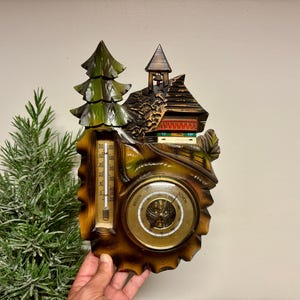 May include: A wall-mounted weather station featuring a carved wooden chalet and a green pine tree. The device includes a vertical thermometer with a scale in degrees Celsius and a circular barometer with a brass-colored dial and German text.