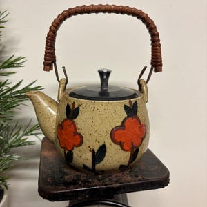 May include: A vintage ceramic teapot with a woven handle and a black lid. The teapot is beige with orange floral designs and black accents. The teapot is sitting on a black scale.