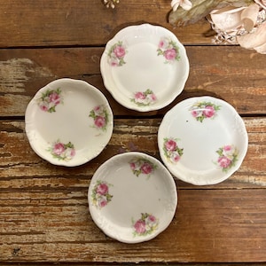 May include: Four small, round white porcelain saucers with scalloped edges, each decorated with a delicate floral pattern featuring pink and white roses with green leaves arranged in clusters around the inner rim of the dishes.