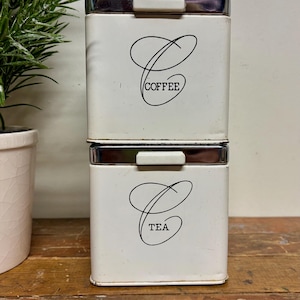 May include: Two white rectangular canisters with silver trim and a handle on top. The top canister reads "COFFEE" in black script, and the bottom canister reads "TEA". The canisters are stacked on top of each other.