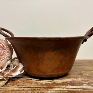 May include: A vintage copper bowl with two handles. The bowl is brown and has a rustic patina. It is sitting on a wooden surface.