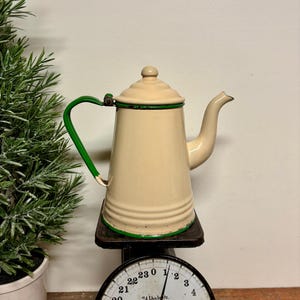 May include: Cream-colored enamelware coffee pot with a green handle and trim, sitting on a vintage black scale. The scale has a round face with black numbers and a needle pointing to the weight. A small green plant is visible on the left.
