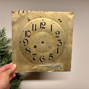 May include: Antique clock face with black numerals on a gold-toned square metal plate. The clock face features the numbers 1 through 12, with two holes in the center. The metal plate has a distressed appearance.
