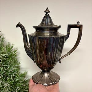 May include: A silver-colored antique teapot with a fluted body, a curved spout, and a square handle. The teapot has a decorative lid with a finial and sits on a scalloped base. The teapot is held in a person's hand.