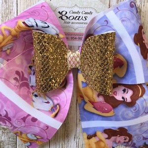 Puede incluir: Un gran lazo de brillantina dorada con un estampado de tela de Belle y la Bestia rosa y azul. El lazo está atado con una cinta dorada y tiene un pequeño lazo de malla dorada en el centro. El lazo está etiquetado como "Candy Candy Bows" y "Hair Accessories".